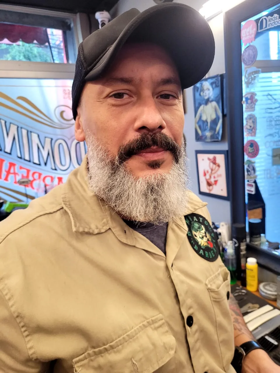 Fabio, barbeiro da Dominato, de boné e camisa cáqui com o emblema da barbearia, no salão.