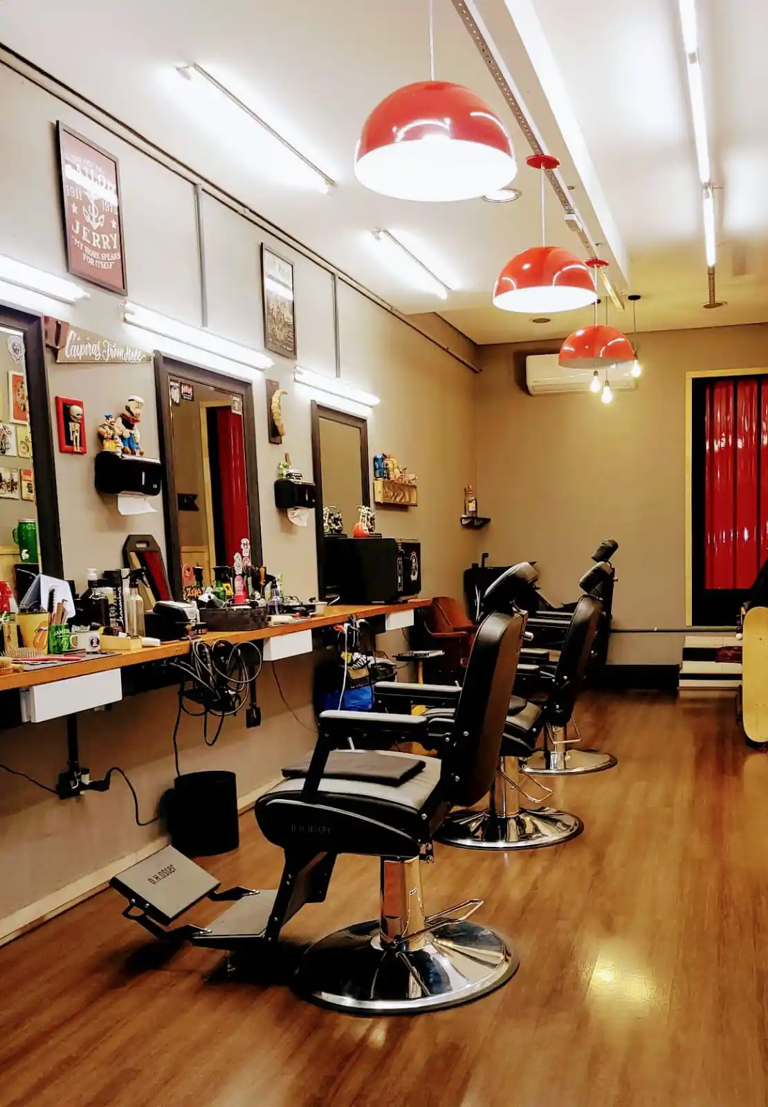 Salão da barbearia com cadeiras de barbeiro clássicas, luminárias vermelhas e piso de madeira.