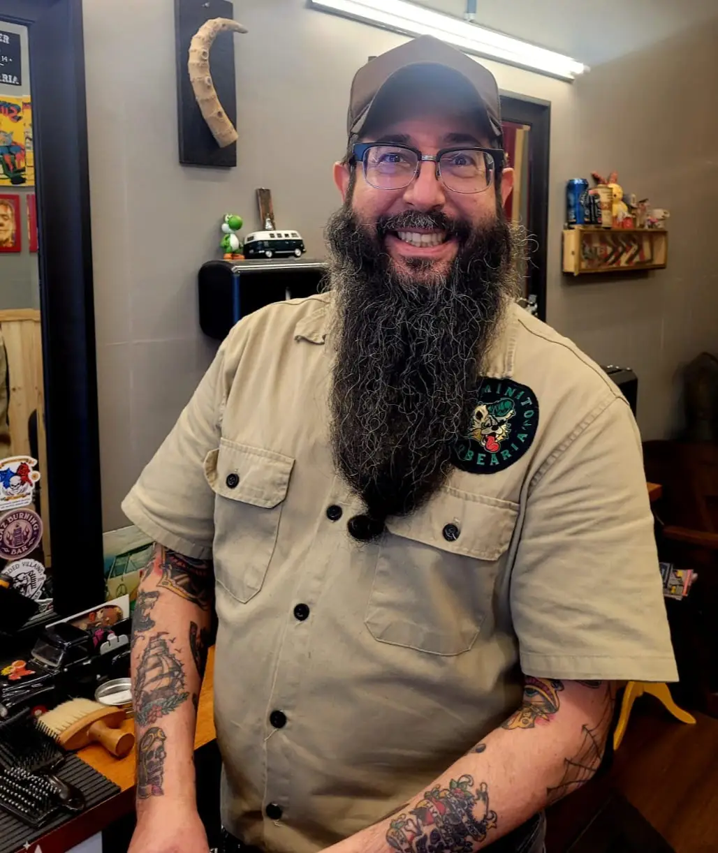 Luizão, barbeiro da Dominato, de barba longa e camisa da barbearia, sorrindo no salão.