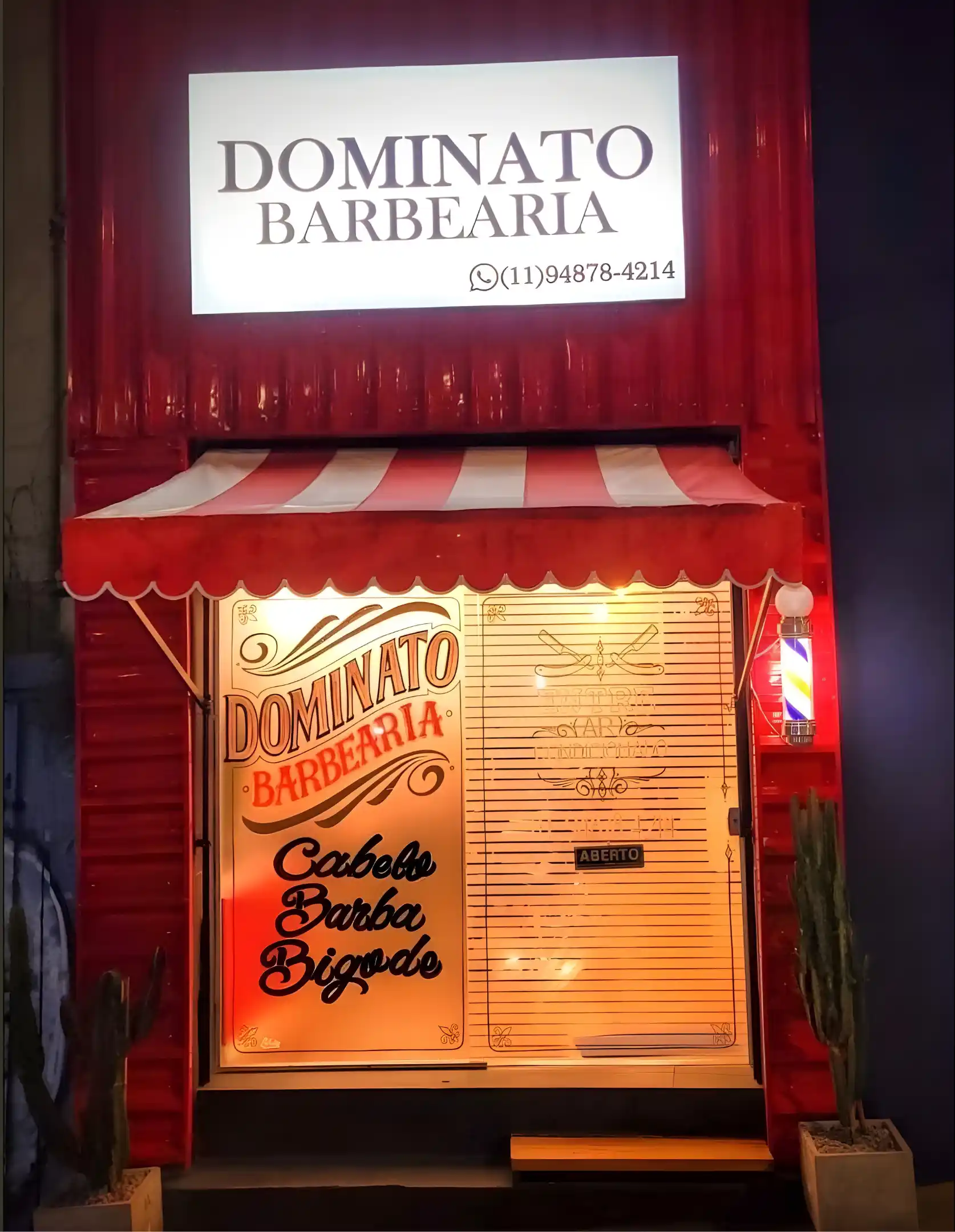 Fachada vermelha da Dominato Barbearia à noite, com letreiro iluminado, toldo listrado e poste de barbeiro aceso.